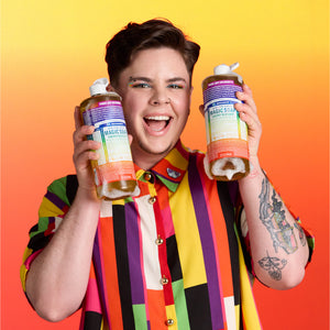 Person holding two bottles of Dr. Bronner's limited-edition Pride Cherry Blossom Magic Soap against a colourful gradient background