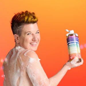 Person with soap suds on a bright orange background holding a bottle of Dr. Bronner's limited-edition Cherry Blossom Pride Magic Soap.
