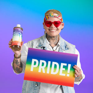 Person holding a bottle of Dr. Bronner's limited-edition Cherry Blossom Pride Soap and a rainbow-colored 'PRIDE!' sign against a colourful gradient background